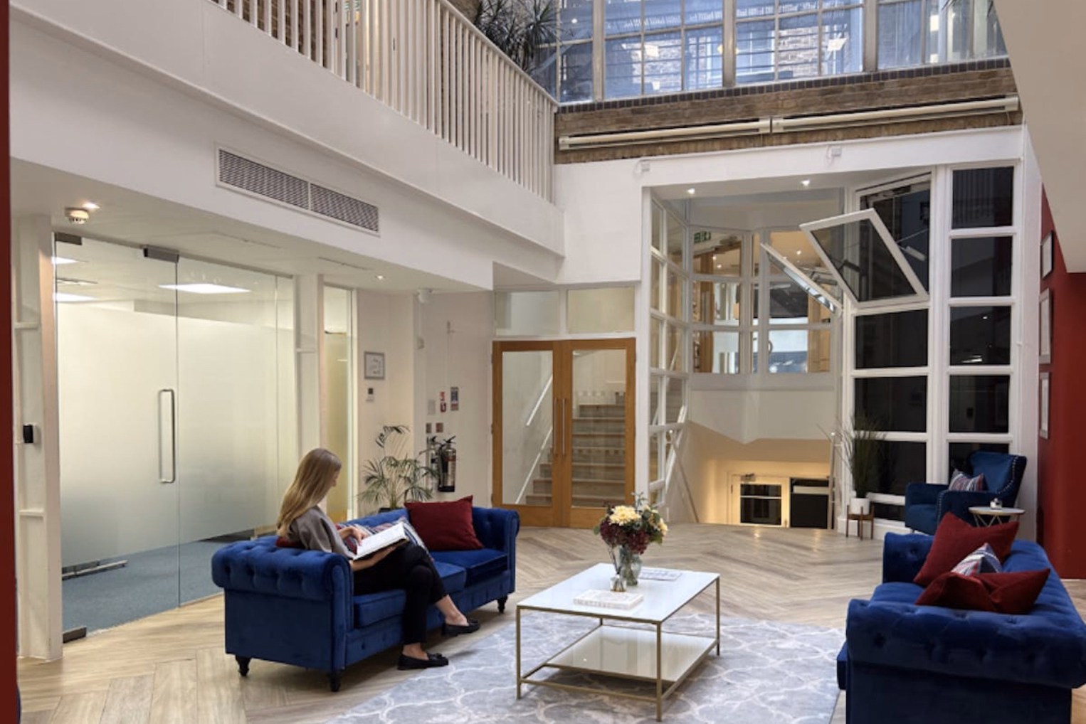 Blue velvet sofas with red throw pillows frame a white table on a patterned rug in a multi-level atrium office lobby, featuring glass partitions and an upper-level balcony.