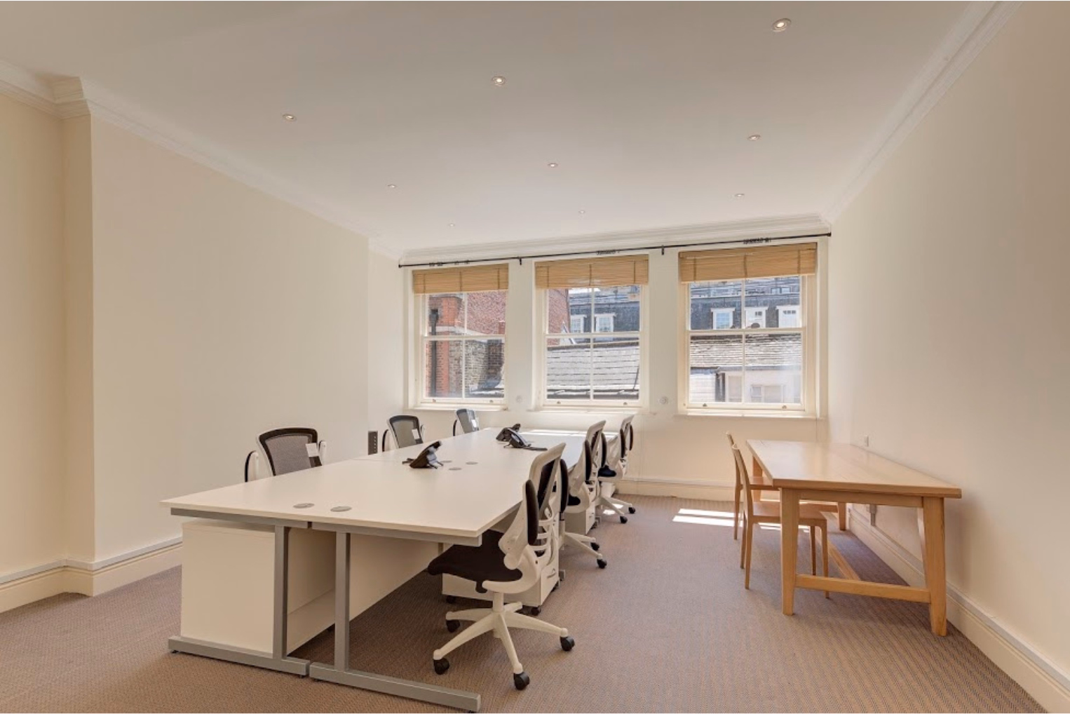 Office chairs surround a white desk island with under-desk storage, set beside a separate wooden side table with matching chairs in an office with sash windows and blinds.