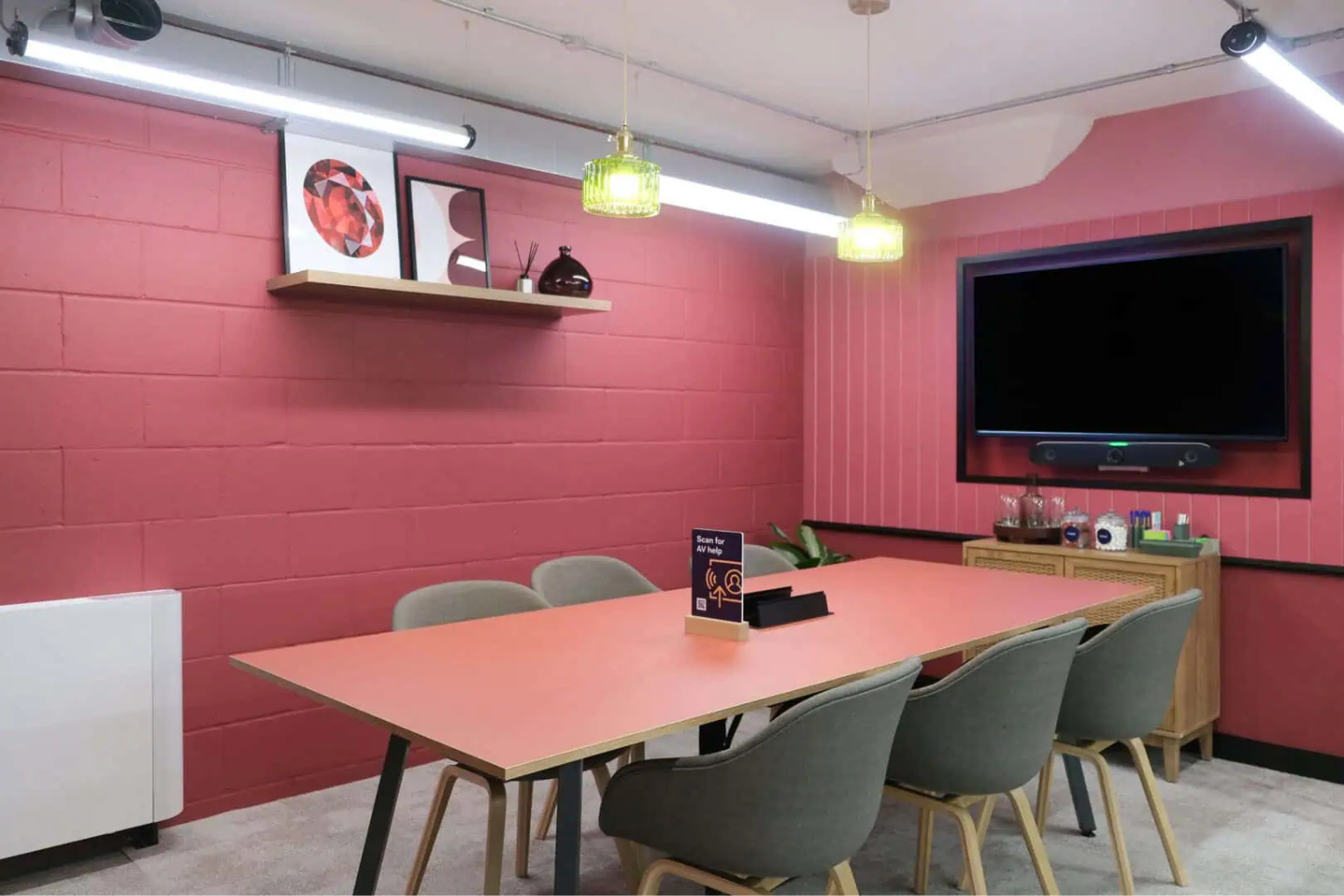 A vibrant meeting room with pink walls, a long coral-coloured table, grey chairs, and a wall-mounted television.