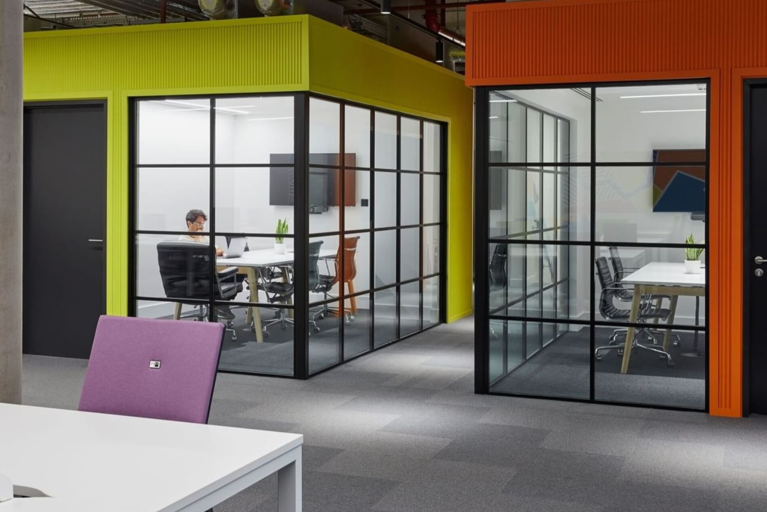 Individuals work at white desks inside glass-partitioned private offices within a modern, colourful workspace.
