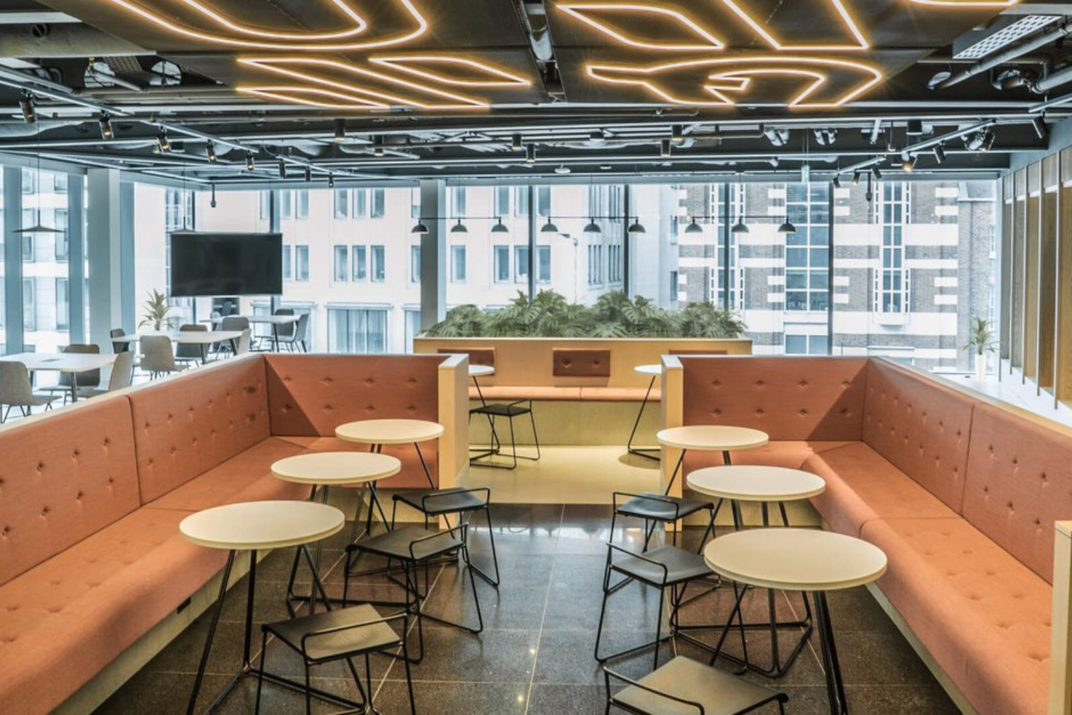 An office canteen featuring curved peach-coloured seating and small round tables under decorative linear ceiling lights.