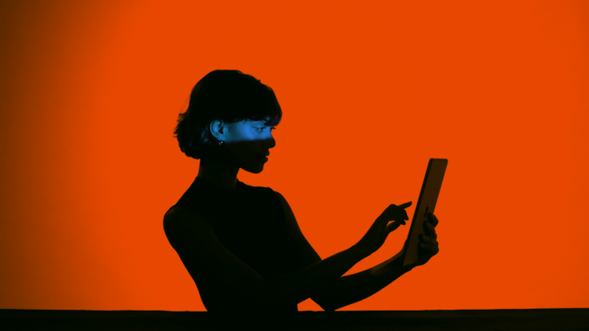 Silhouette of a woman holding and using a tablet, lit by blue light against a bright orange background.
