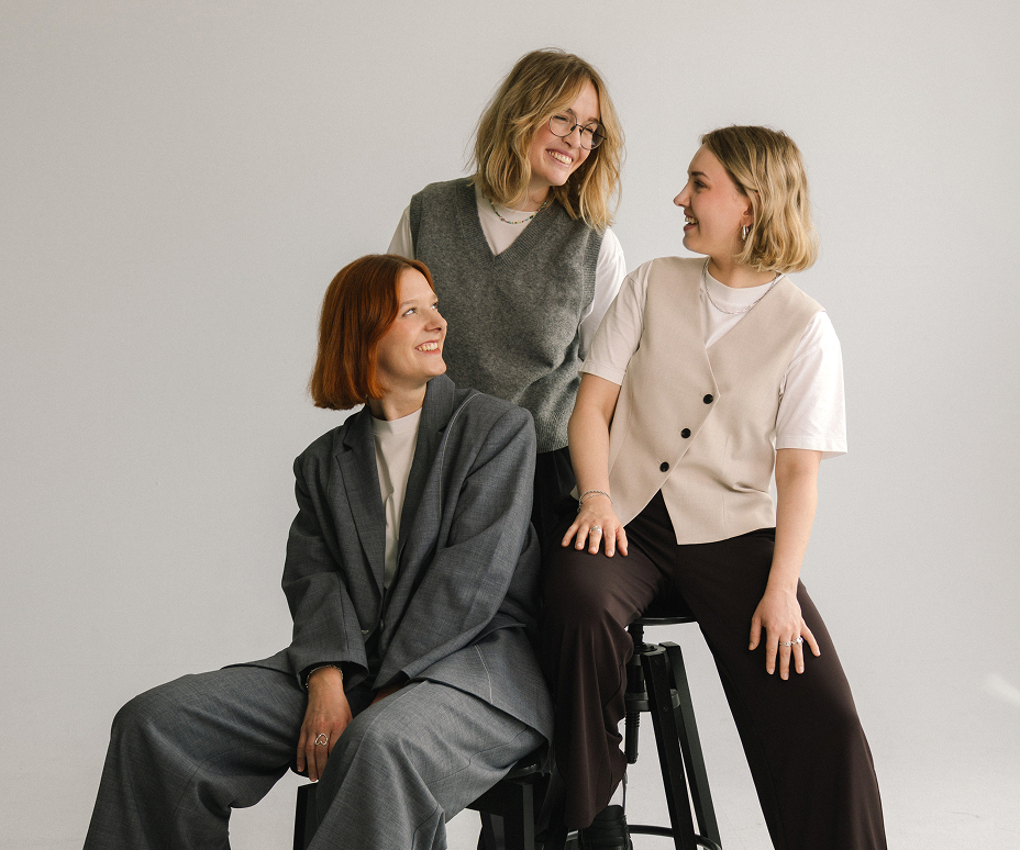 Three women from Studio Tülü posing together in a studio setting, dressed in modern business casual outfits.