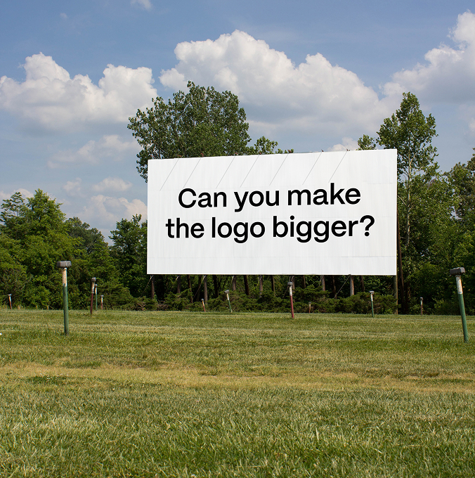 Large outdoor billboard on a grassy field with the text “Can you make the logo bigger?” in bold black letters.