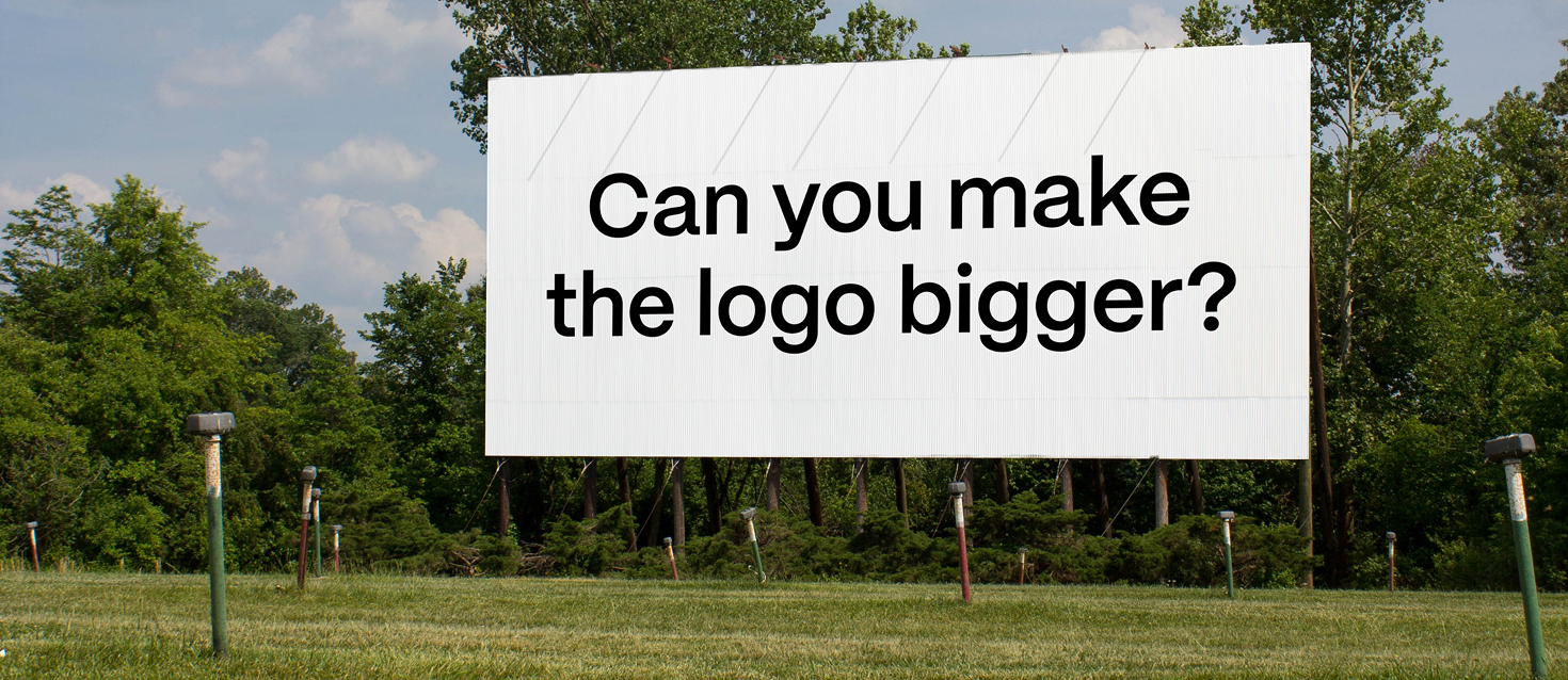 Large outdoor billboard on a grassy field with the text “Can you make the logo bigger?” in bold black letters.
