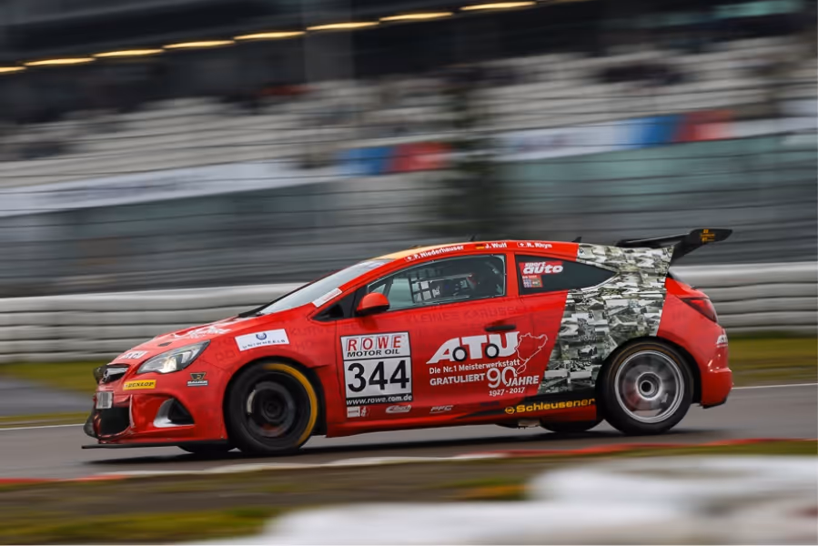 Red race car number 344 with sponsor logos including ROWE Motor Oil on a track with blurred background showing motion.
