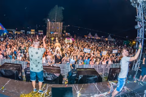 Two performers on stage facing a large, energetic crowd at an outdoor night concert.