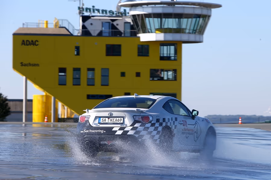 White sports car with checkered pattern drifting on wet track in front of a yellow control tower at Sachsenring race circuit.