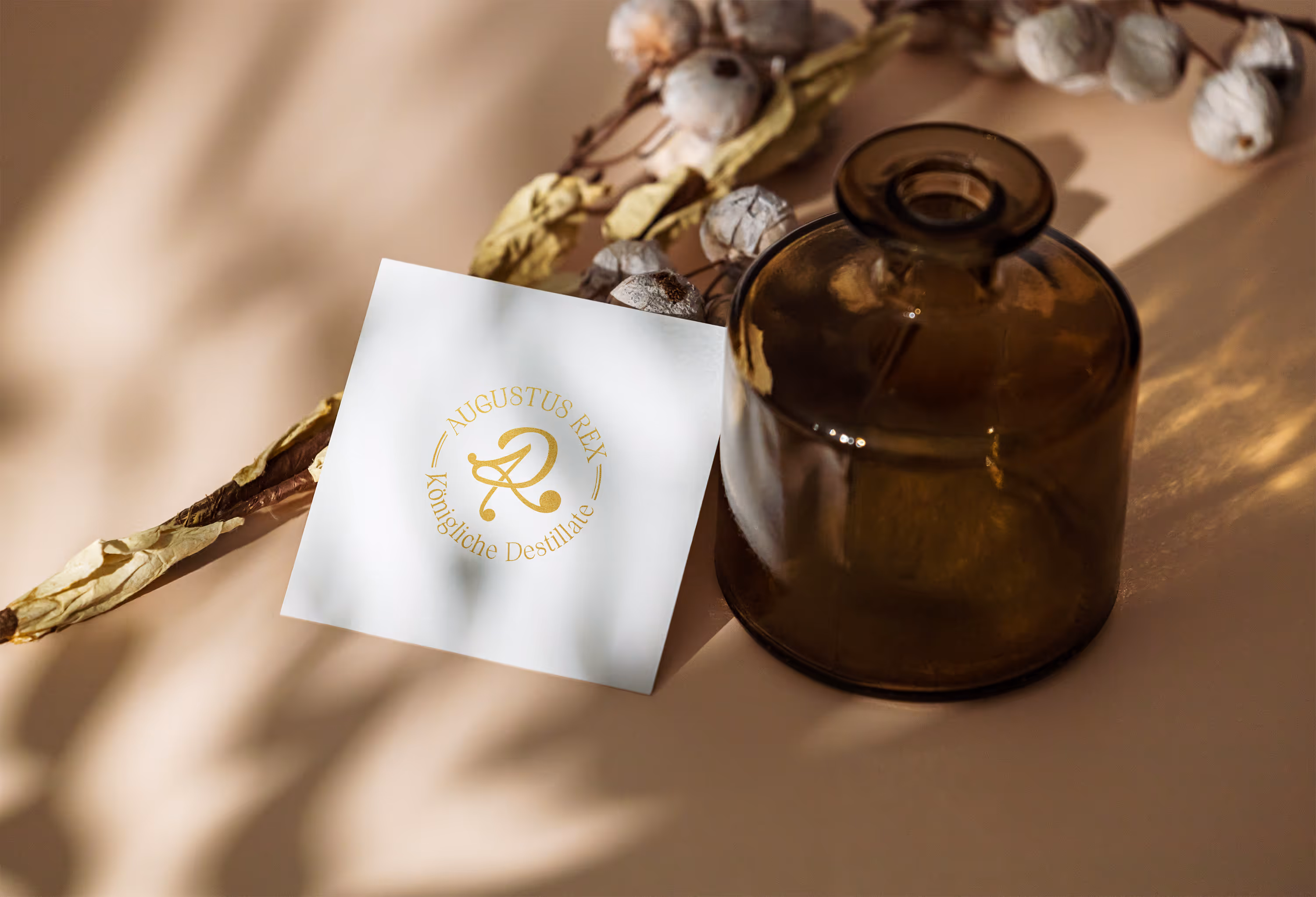 Amber glass bottle next to a white card with a gold logo reading 'Augustus Rex Königliche Destillate' on a beige surface with dried cotton stems.