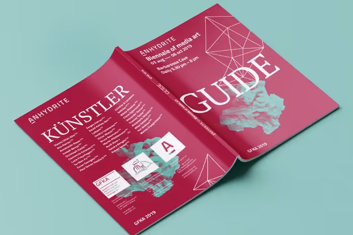 Open magenta guidebook for Anhydrite Biennale of Media Art 2019 featuring abstract geometric and cloud-like shapes.
