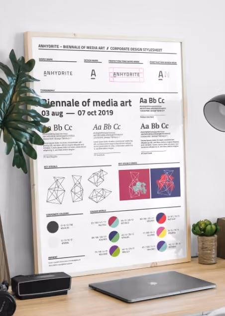Framed corporate design stylesheet poster on a desk next to a laptop and small plant, featuring fonts, typography, geometry diagrams, and color palettes for Anhydrite Biennale of Media Art 2019.