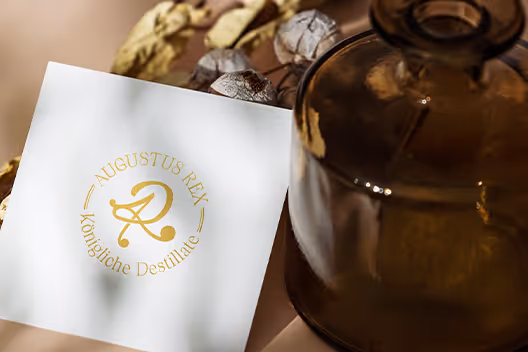 White card with golden logo and text 'Augustus Rex Königliche Destillate' next to a brown glass bottle and dried flowers on a neutral surface.