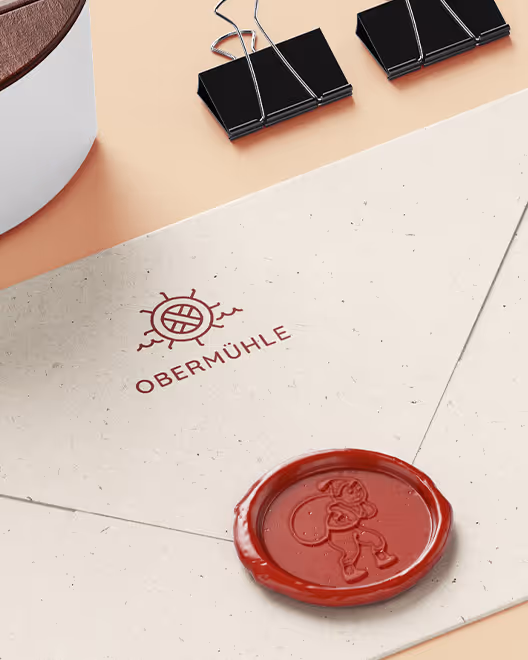 Envelope with Obermühle logo and a red wax seal featuring a character, placed on a beige surface alongside two black binder clips and part of a white container with a wooden lid.
