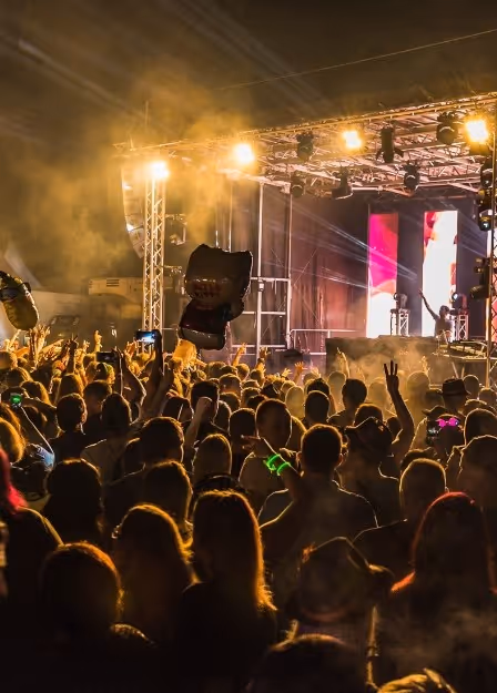 Crowd of people enjoying an outdoor concert at night with bright stage lights and some holding up phones and balloons.