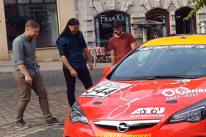 Three men standing on a cobblestone street next to a red and yellow race car with the Opel logo and various sponsor decals.