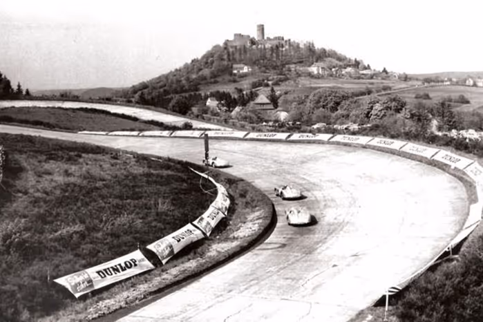 Black and white photo of a vintage car race on a curved concrete racetrack with a hilltop castle in the background.