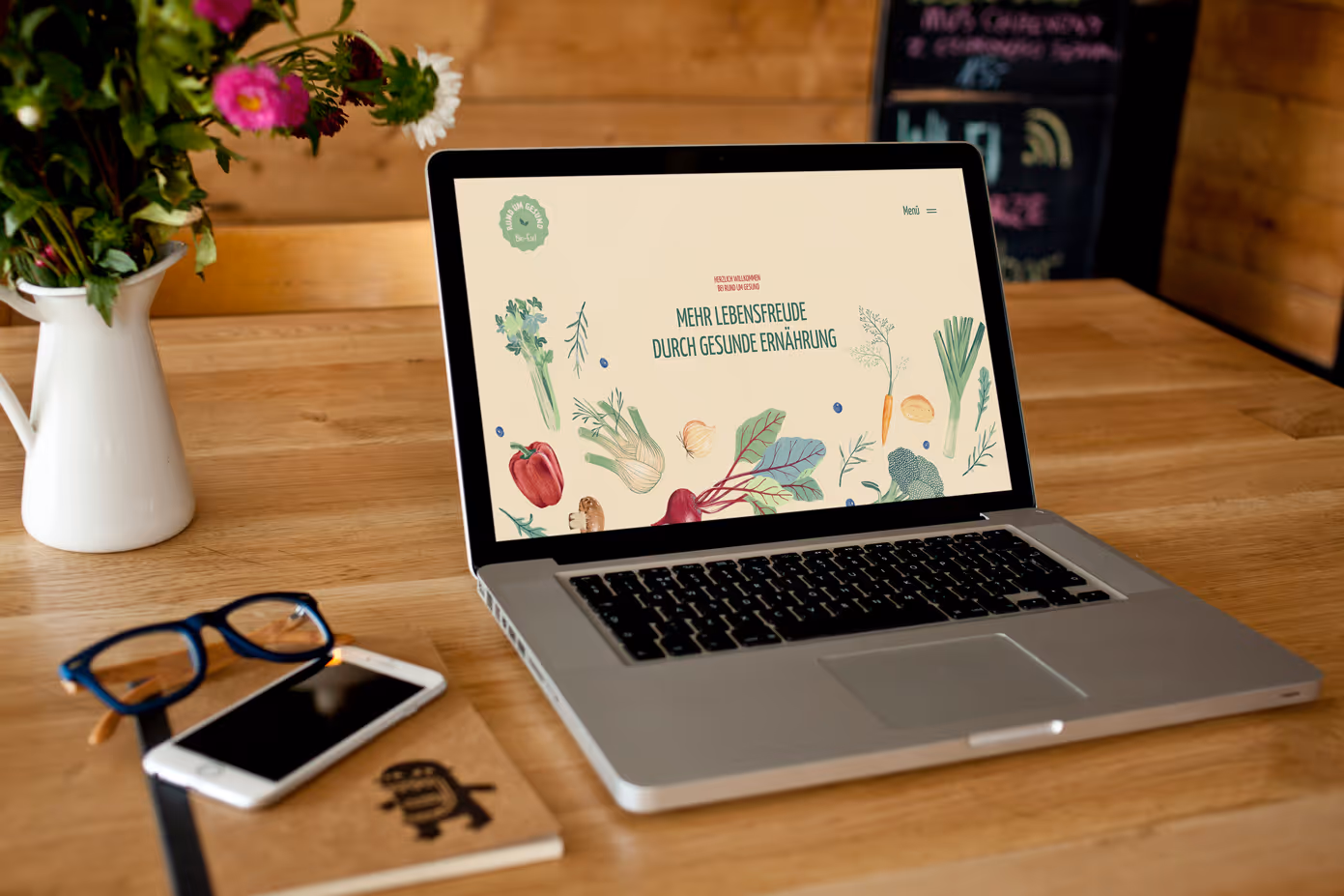 Laptop on wooden desk displaying a website with the text 'Mehr Lebensfreude durch gesunde Ernährung' surrounded by illustrations of vegetables.