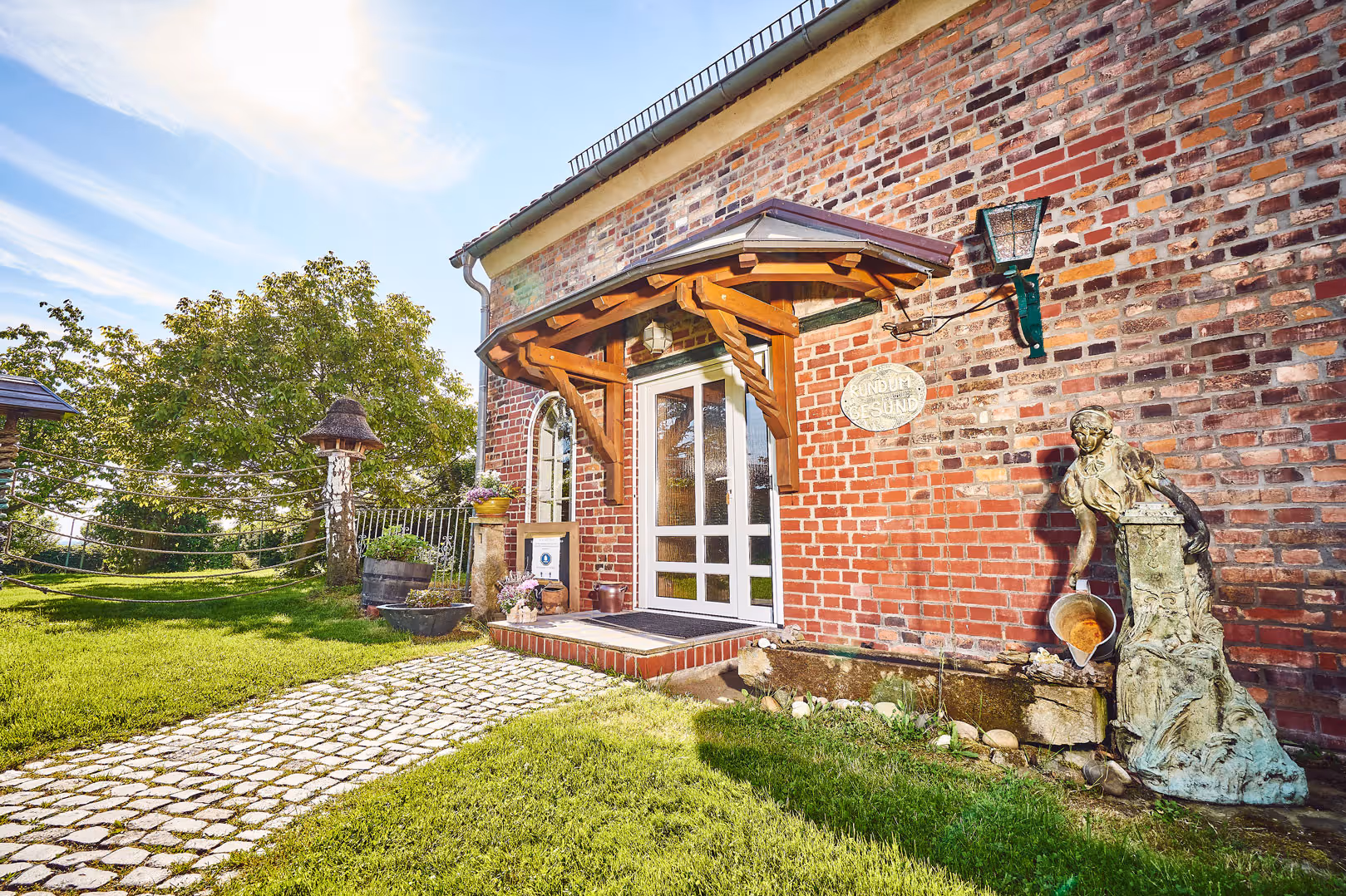 Entrance of a brick building with a wooden canopy, a cobblestone path, and a decorative statue pouring water on the right side.