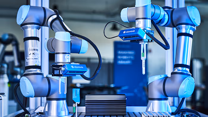 Two silver and blue robotic arms in a laboratory or industrial setting with 'robominds' branding on attached sensors.