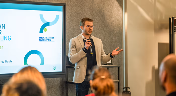 Man in beige blazer and glasses speaking into a microphone during a presentation in front of an audience.