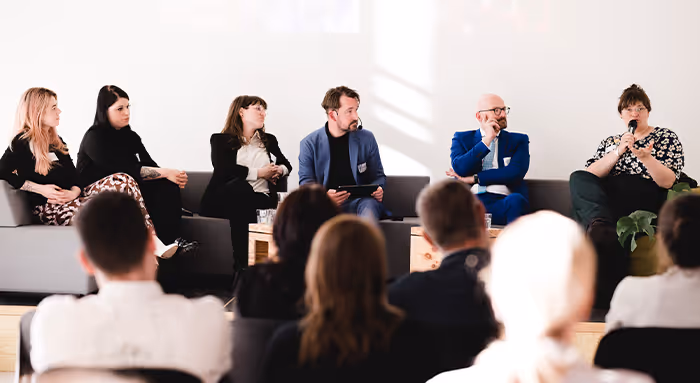 Panel of six people seated on a stage, one woman speaking into a microphone, audience in foreground.