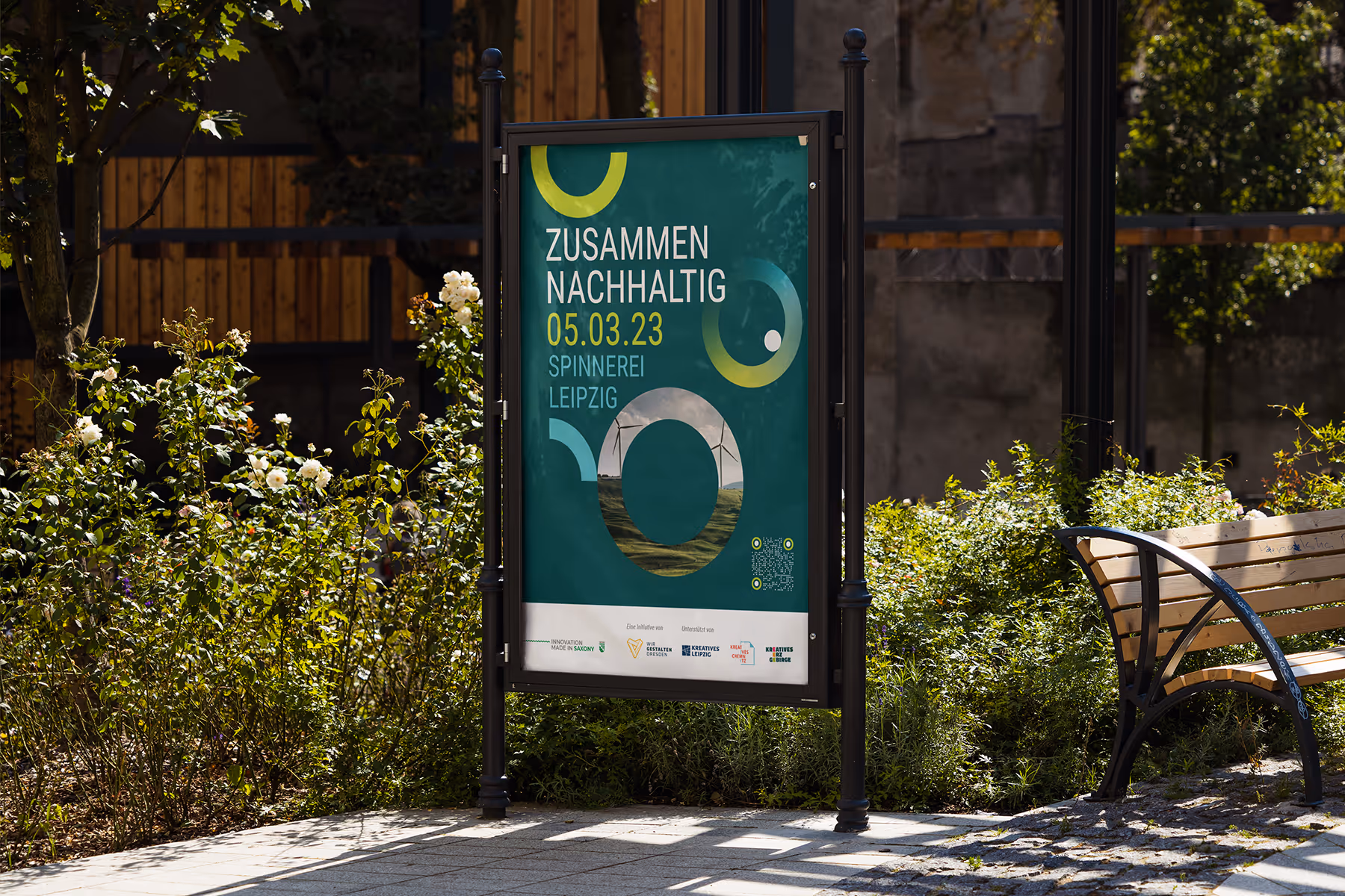 Outdoor poster in a black frame promoting a sustainability event in Leipzig on March 5, 2023, surrounded by plants and a wooden bench.