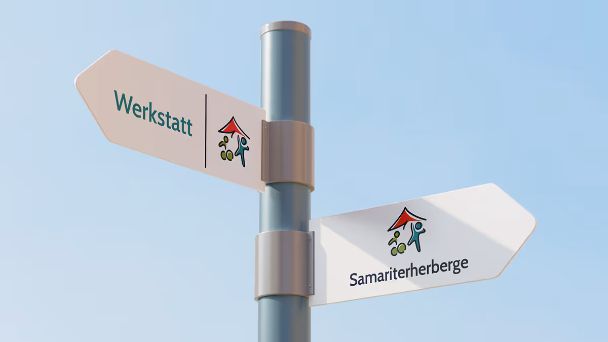 Two white directional signs on a pole against a clear blue sky, one pointing left labeled 'Werkstatt' and the other right labeled 'Samariterherberge,' both with small logos featuring a red roof and green and blue figures.