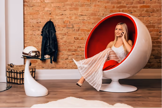 Young woman in white dress sitting in a modern white and red egg chair against a brick wall, talking on a phone.