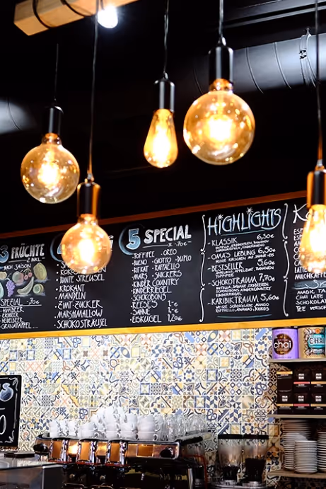 Warm glowing vintage-style pendant lights hanging above a café counter with a patterned tile backsplash and a chalkboard menu.