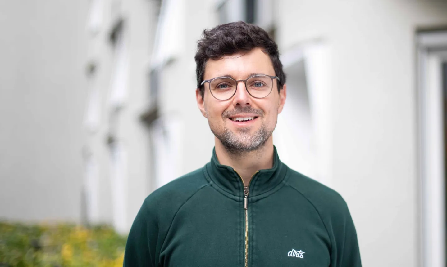 Ein lächelnder Mann mit kurzen dunklen Haaren und Brille trägt einen grünen Reißverschluss-Pullover vor einem unscharfen weißen Hintergrund.