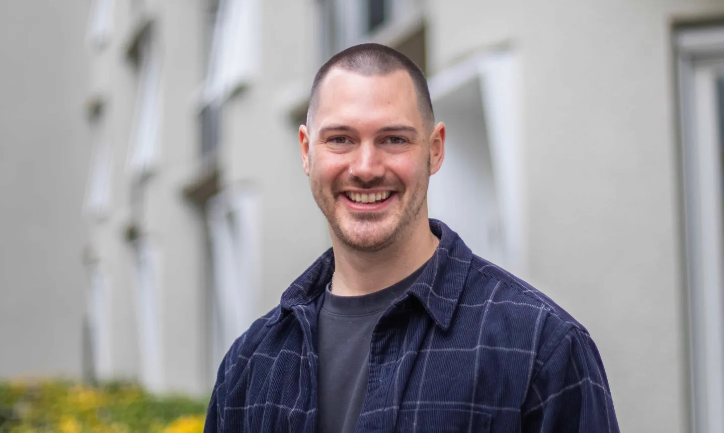 Junger Mann mit kurzem Haar und Bart, der in einem karierten dunkelblauen Hemd und schwarzem T-Shirt lächelt.