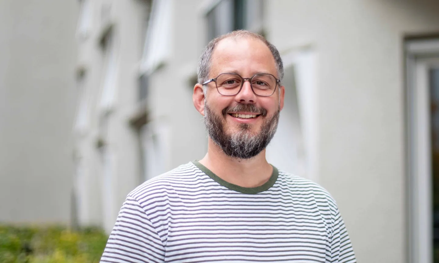Ein lächelnder Mann mit Brille, Bart und gestreiftem T-Shirt vor einem verschwommenen Gebäudehintergrund.