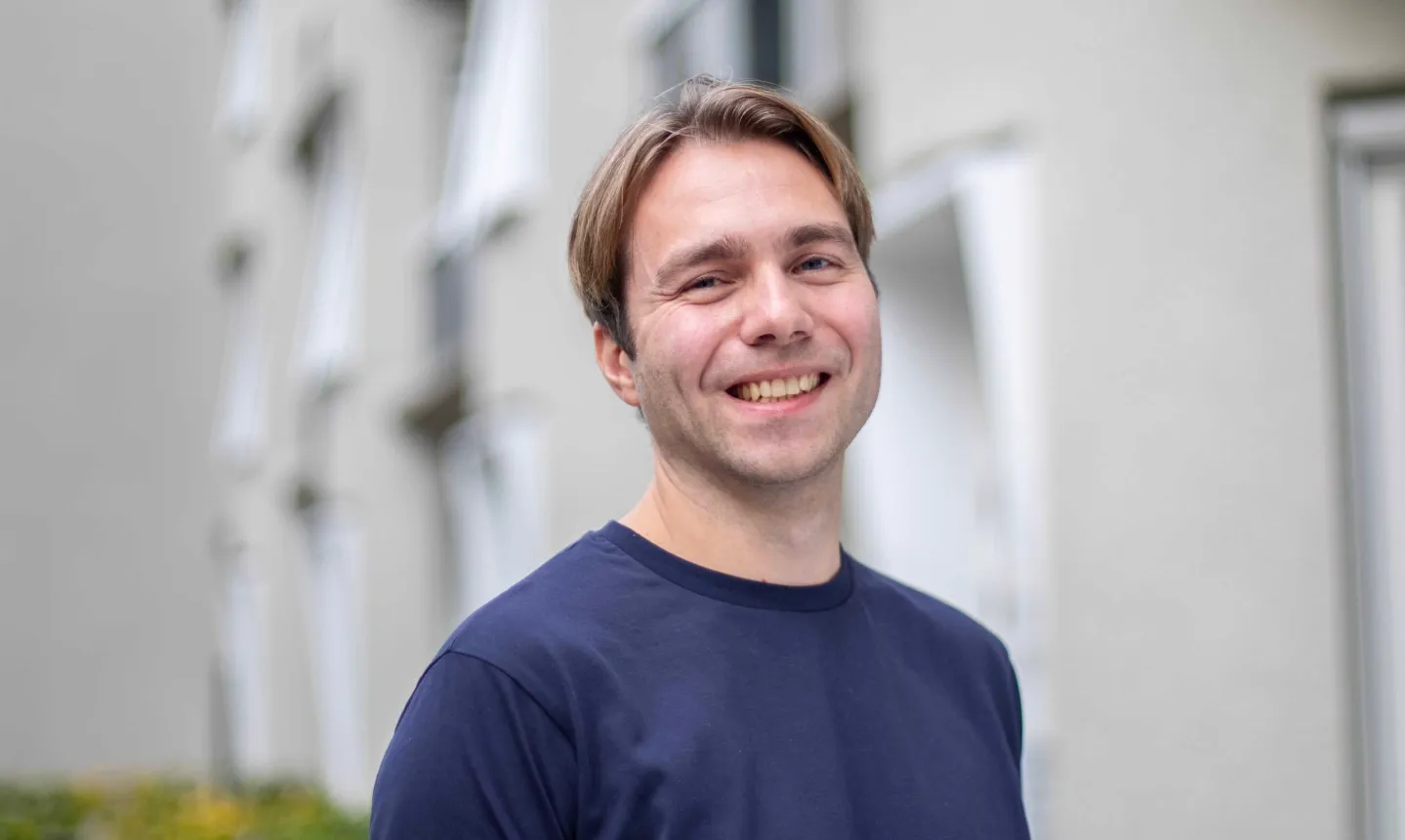 Ein lächelnder Mann mit kurzen, braunen Haaren trägt ein dunkelblaues T-Shirt vor einem unscharfen, hellen Hintergrund.