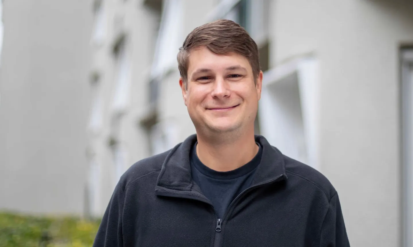 Lächelnder Mann mit braunen Haaren und dunkler Jacke vor verschwommenem Gebäude.