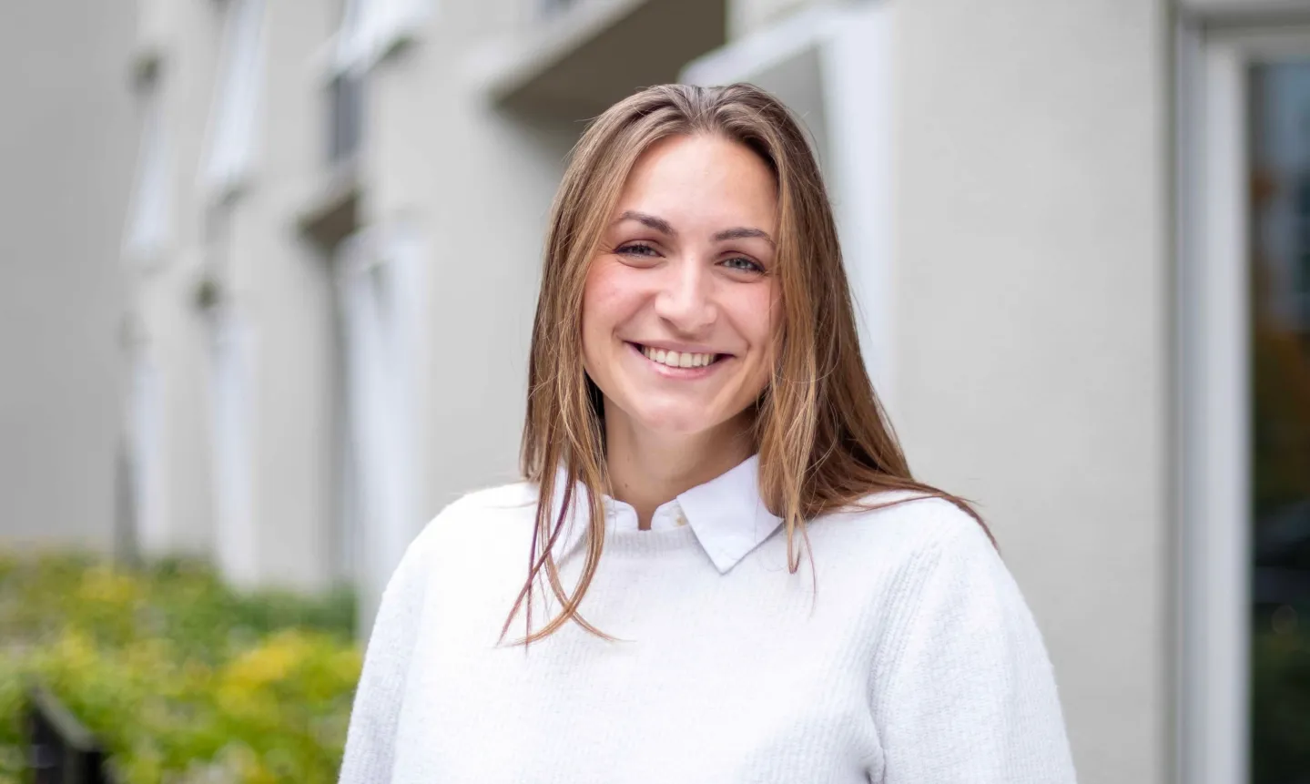 Porträt einer lächelnden Frau mit langen braunen Haaren und weißem Pullover vor einem Gebäude.