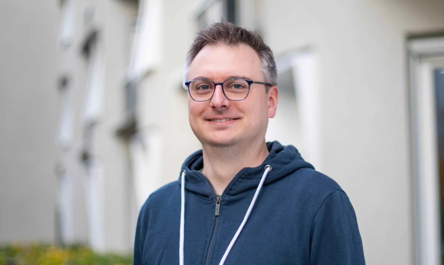 Mann mit Brille und dunkelblauem Hoodie lächelt vor verschwommenem Gebäudehintergrund.