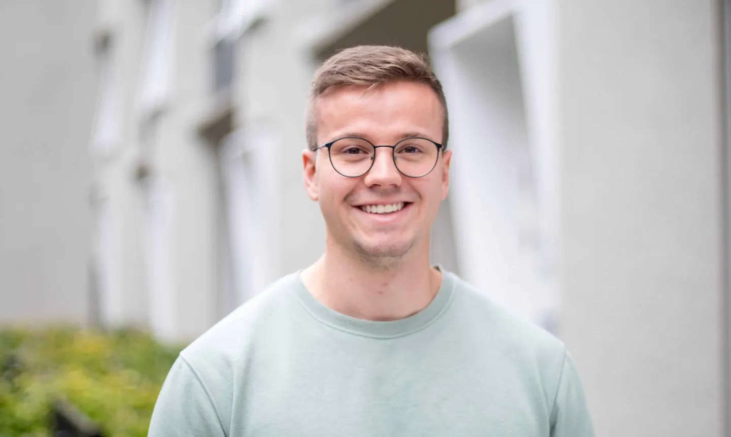 Junger Mann mit kurzen hellbraunen Haaren, der eine Brille trägt und in einem hellgrünen Pullover vor verschwommenem urbanem Hintergrund lächelt.