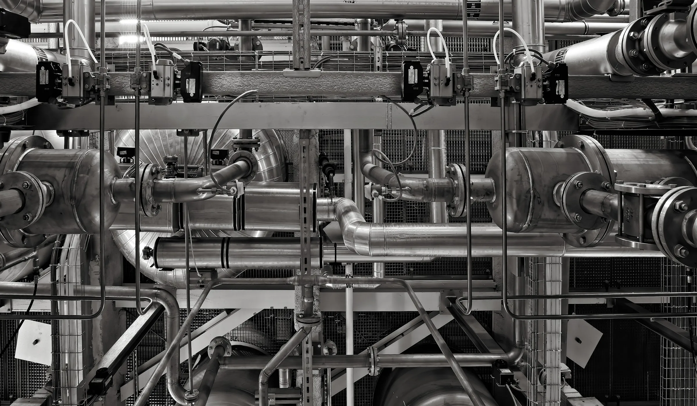 Schwarz-weißes Bild eines komplexen Netzwerks aus Metallrohren, Ventilen und Verbindungsstücken in einer Industrieanlage oder Fabrik.