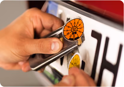 Nahaufnahme: Eine Hand befestigt mit einem Werkzeug eine orangefarbene Prüfplakette auf einem Kennzeichen.