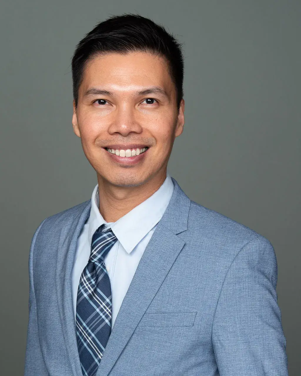 Smiling man in a light blue suit jacket, white shirt, and plaid tie against a gray background.