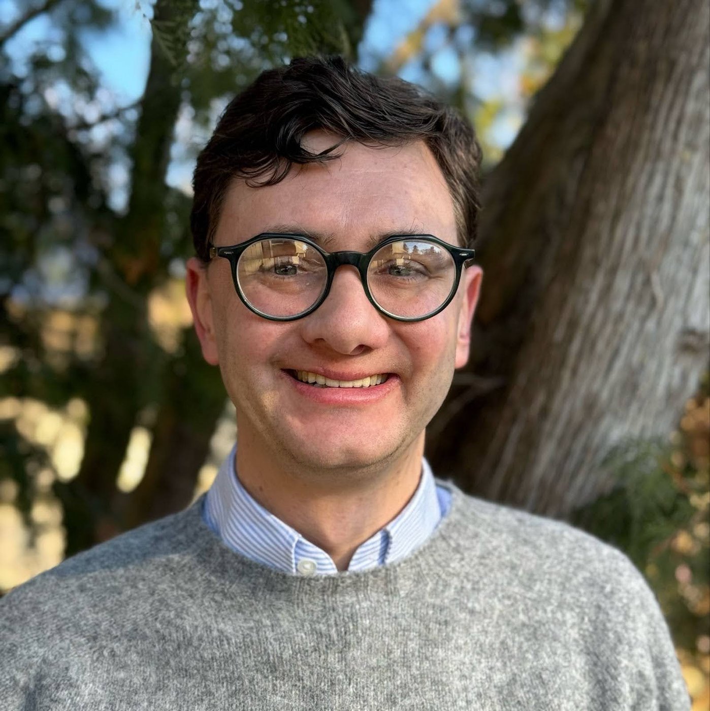Smiling man with dark hair wearing round glasses, a striped shirt, and a gray sweater outdoors.