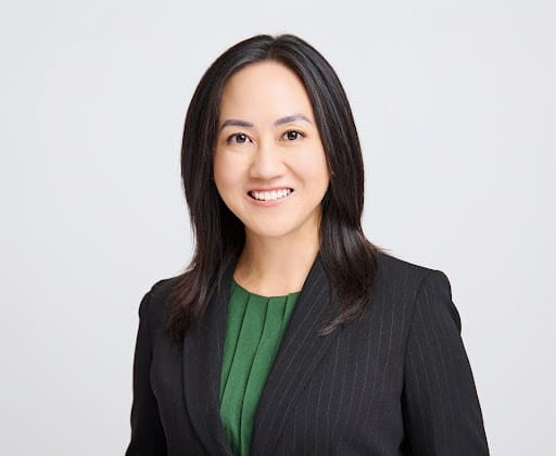 Smiling woman with straight black hair wearing a dark blazer and green top on a light background.