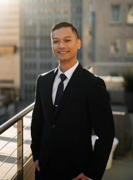 Young man in a black suit and tie smiling outdoors with city buildings in the background.