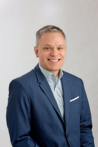 Smiling man with short hair wearing a blue suit jacket and light blue shirt against a plain background.