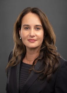 Professional headshot of a woman with long brown hair wearing a dark blazer and pearl earrings.