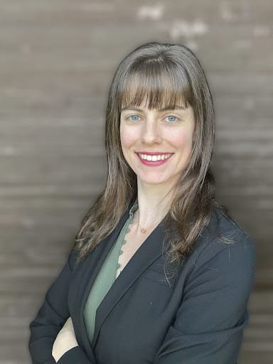 Smiling woman with long brown hair and bangs wearing black blazer and green top, arms crossed.