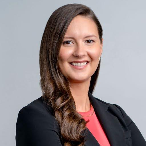 Smiling woman with long dark hair wearing a black blazer and coral top against a gray background.