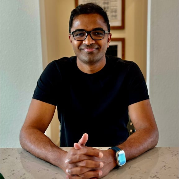 Smiling man wearing glasses and a black shirt sitting at a table with clasped hands and a smartwatch.