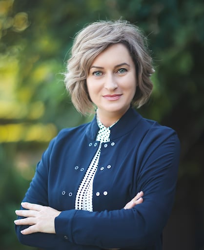 Woman with short curly blonde hair wearing a blue jacket and white polka-dot shirt, smiling with arms crossed.
