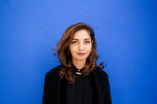 Woman with wavy brown hair in black clothing standing against a bright blue wall.